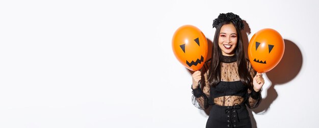 Image of cheerful asian woman in witch costume celebrating halloween holding balloons with scary fac