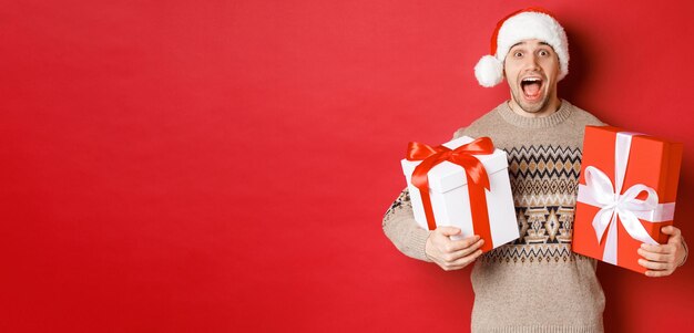 Изображение веселого привлекательного парня с рождественскими подарками, стоящего в новогодней шапке и зимнем свитере, изумленно улыбающегося, стоящего на красном фоне.