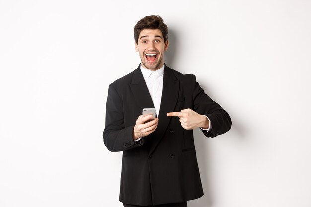 Image of cheerful businessman looking amazed, pointing at mobile phone, standing in suit over white background