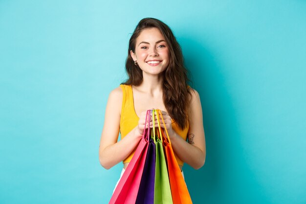 Изображение веселой девушки, идущей по магазинам, держа сумки с покупками и улыбаясь в камеру, стоя на синем фоне.