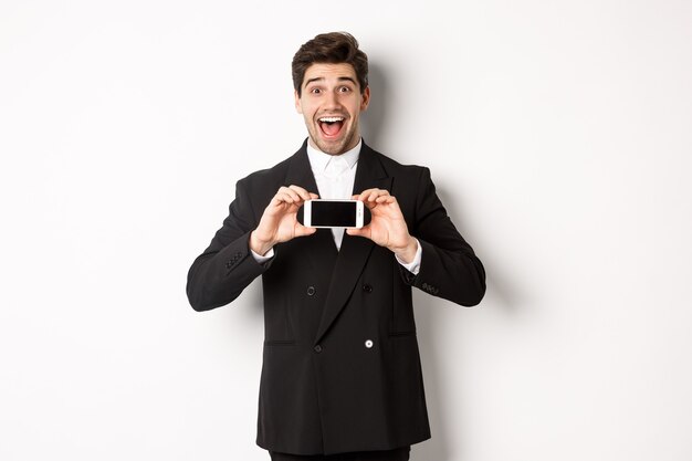 Image of cheerful, handsome man in black suit, showing smarthone screen and looking amazed, standing against white background.