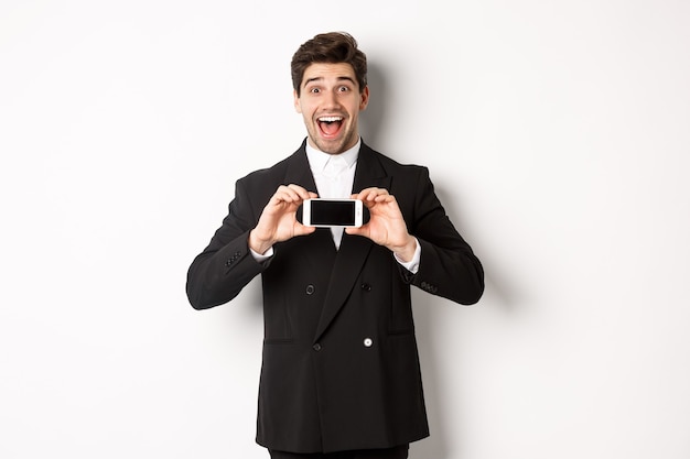 Изображение веселого, красивого мужчины в черном костюме, демонстрирующего экран смартфона и выглядящего изумленно, стоящего на белом фоне