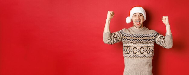 Image of cheerful handsome man in swearer and santa hat celebrating new year triumphing or winning something raising hands up and shouting for joy standing over red background