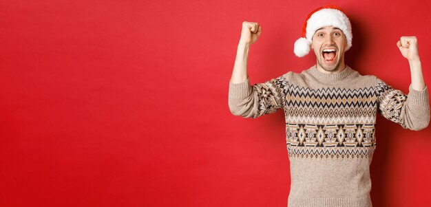 Изображение веселого красивого мужчины в ругательстве и шляпе Санты, празднующего новый год, торжествующего или выигрывающего что-то, поднимающего руки вверх и кричащего от радости, стоящего на красном фоне.