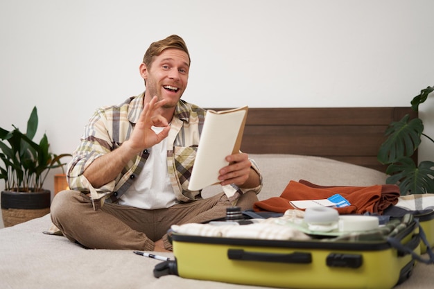 Image of cheerful man shows okay sign packs clothes in suitcase goes on holiday or business trip