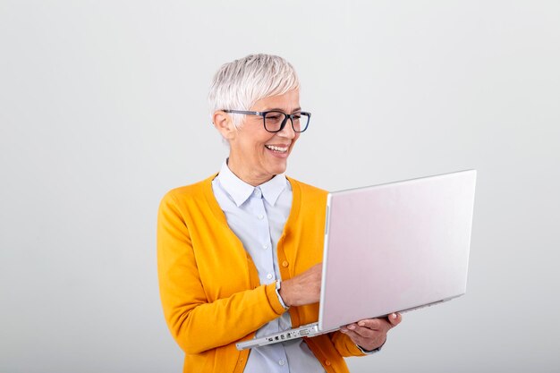 ラップトップコンピューターを使用して灰色の背景の上に孤立して立っている陽気な成熟した女性の画像ラップトップコンピューターを保持している笑顔のシニア女性の肖像画