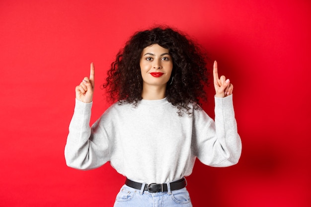 巻き毛の髪型と赤い唇、空のスペースで指を上向き、広告を表示、スタジオの背景にカジュアルな服を着て立っている陽気な笑顔の女性の画像