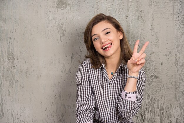 Изображение веселой женщины в повседневной одежде, улыбающейся и показывающей знак мира двумя пальцами