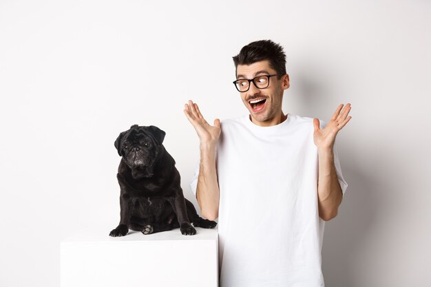 Image of cheerful young man rejoicing, looking at cute black dog pug and smiling, standing over white background.