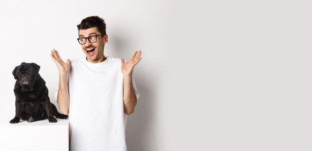 Free photo image of cheerful young man rejoicing looking at cute black dog pug and smiling standing over white