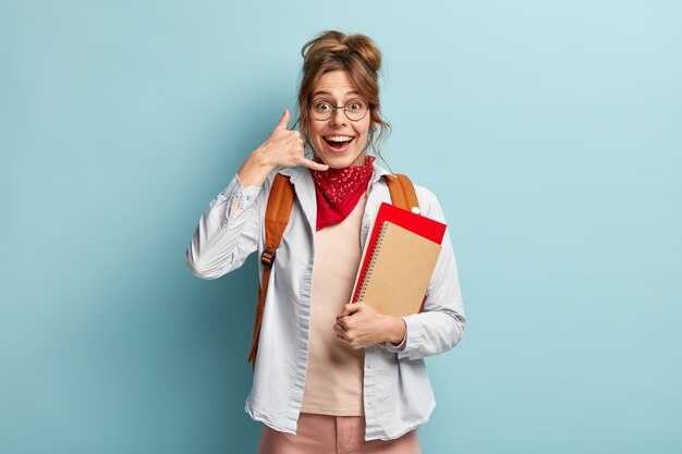 Image of cheerful young woman makes phone gesture, wears shirt and red bandana