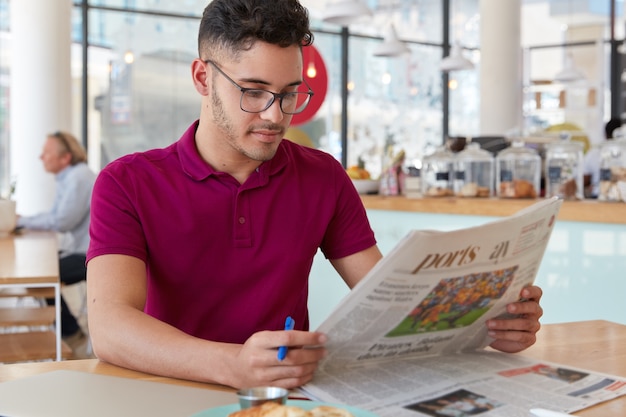 Image of concentrated man with serious facial expression, reads newspaper, finds out world news, holds pen to underline main facts, wears glasses and casual t shirt, poses over coffee shop interior