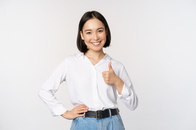 Image of confident asian woman showing thumb up in approval recommending like smth good standing over white background