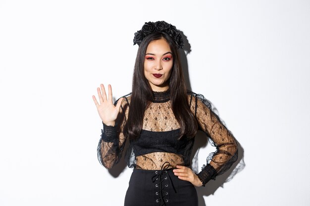 Изображение уверенно красивой азиатской женщины в костюме хеллоуина показывает пять пальцев, поднимая руку, чтобы поздороваться, стоя над белой предпосылкой.