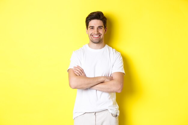 Image of confident caucasian man smiling pleased, holding hands crossed on chest and looking satisfied, standing over yellow background.