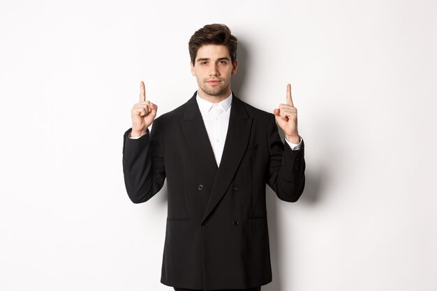 Image of confident and handsome man in formal suit, pointing fingers up, showing copy space on white background