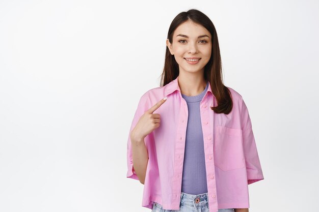 Image of confident young woman pointing finger at herself smiling and looking selfassured standing against white background