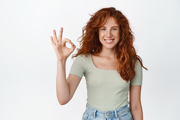 Изображение уверенной в себе молодой женщины с рыжими длинными здоровыми волосами показывает хороший знак и улыбается, довольная, довольная реакция стоит на белом фоне