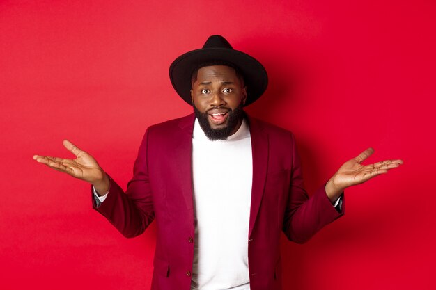 Image of confused Black man asking question, spread hands sideways and staring camera clueless, standing against red background