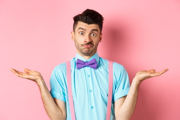 Image of confused guy in bow-tie and suspenders know nothing, shrugging shoulders and looking clueless, standing over pink background.