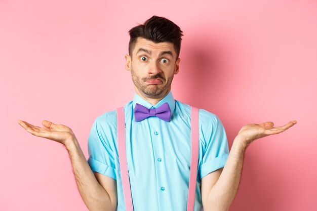 Image of confused guy in bow-tie and suspenders know nothing, shrugging shoulders and looking clueless, standing over pink background.