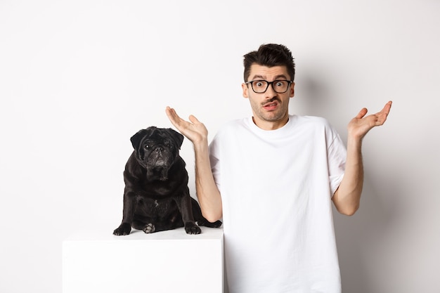 Image of confused man in glasses raising hands and shrugging complicated, standing near black pug dog over white background.