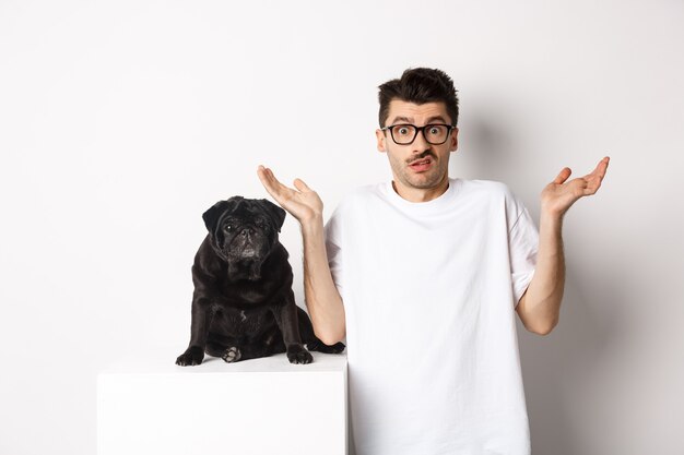 白い背景の上に黒いパグ犬の近くに立って、手を上げて複雑に肩をすくめる眼鏡をかけた混乱した男の画像。