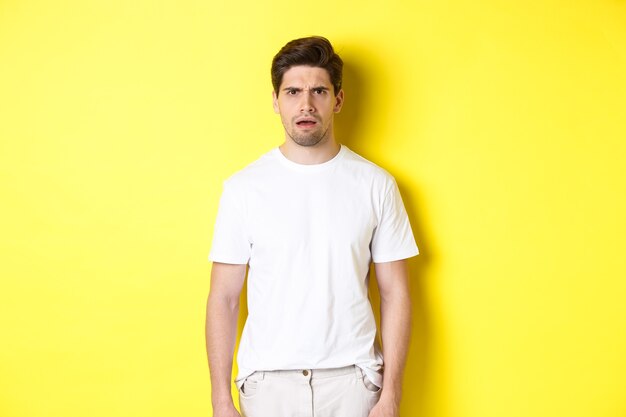 Image of confused and puzzled man cant understand something, frowning and looking shocked, standing over yellow background