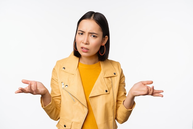 Free photo image of confused young asian woman shrugging shoulders looking puzzled and clueless at camera cant understand standing against white background