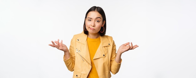 Image of confused young asian woman shrugging shoulders looking puzzled and clueless at camera cant understand standing against white background