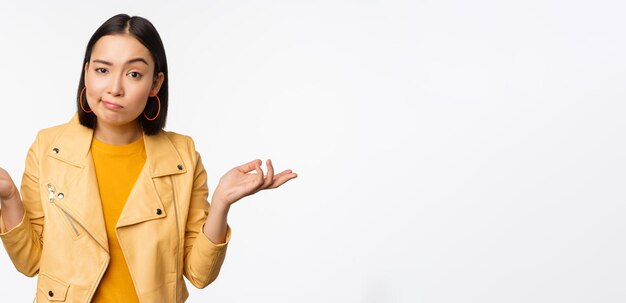 Image of confused young asian woman shrugging shoulders looking puzzled and clueless at camera cant understand standing against white background