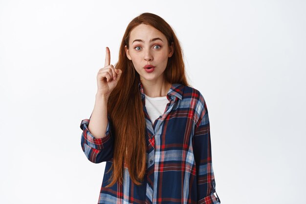 Image of creative ginger teen girl raising finger pointing up pitching an idea found great solution saying plan standing against white background