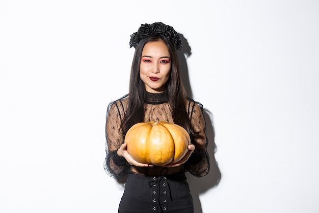 Image of cunning asian woman in black dress, impersonating evil witch on halloween, holding big pumpkin, standing over white background.