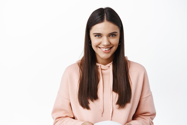 Image of cunning attractive girl smiling looking confident and pensive at camera having plan standing in hoodie against white background Copy space