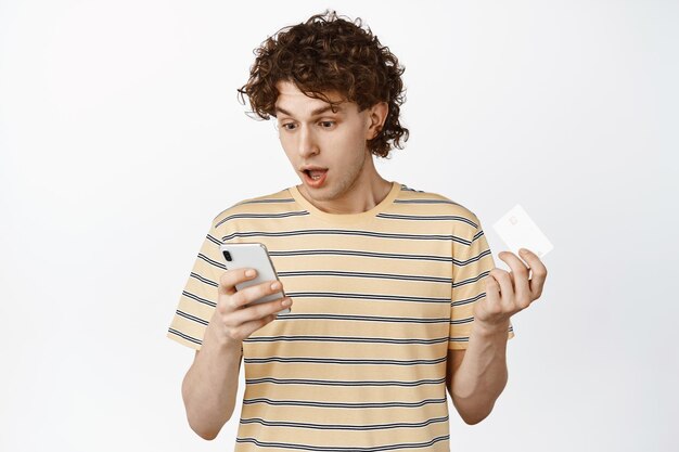 Image of curly guy staring at his phone while placing order holding credit card shopping online app white background