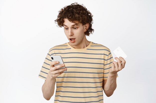 Изображение кудрявого парня, смотрящего на свой телефон во время оформления заказа, держащего онлайн-приложение для покупок кредитной картой на белом фоне