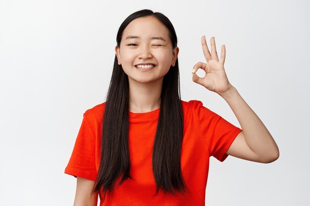 윙크하고 만족스럽게 웃고 있는 귀여운 아시아 여성의 이미지, 승인에 OK 사인을 보여주고, 추천하고, 흰색에 긍정적인 피드백을 남깁니다.