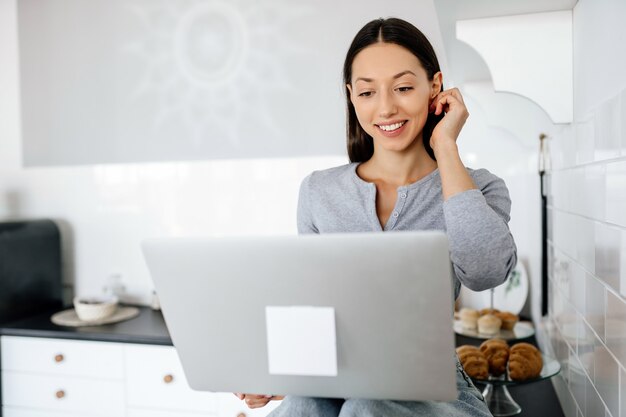 Image of cute brunette woman sitting at kitchen and using laptop