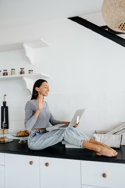 Free photo image of cute brunette woman sitting on the table at kitchen and using laptop