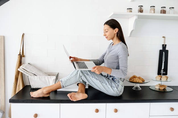 Изображение милой брюнетки женщины, сидящей на столе на кухне и использующей ноутбук