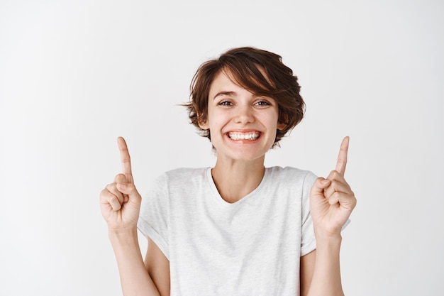 Image of cute and excited woman with short hairstyle and clean glowing skin, pointing fingers up and smiling white wall