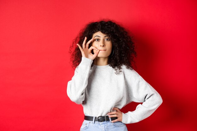 Free photo image of cute girl with curly hair making promise to stay quiet, zipping lips, making seal, hiding a secret, standing dumb on red background