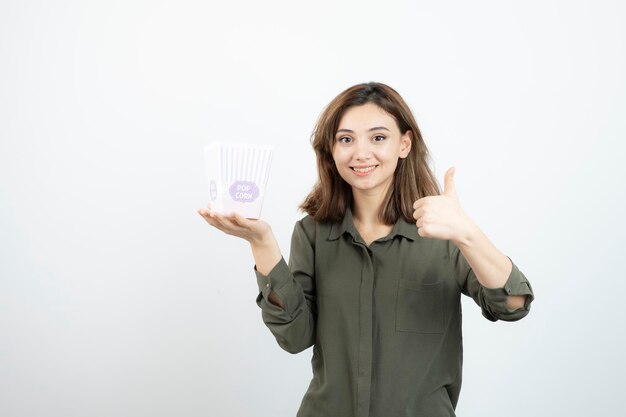 Image of cute girl with popcorn box posing to camera and giving thumbs up. High quality photo