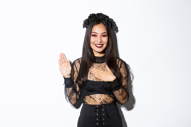 Image of cute sincere asian girl in halloween costume making promise, holding one hand on heart while swearing, being honest, standing over white background.