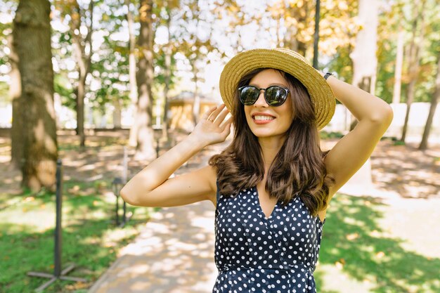 Изображение милой женщины с темными короткими волосами, одетой в платье, гуляет в парке с очаровательной улыбкой. На ней летняя шляпа и черные солнцезащитные очки.