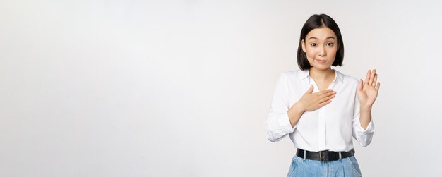 귀여운 젊은 여성 회사원 아시아 여자 학생이 손을 들고 가슴 이름에 손바닥을 대고 흰색 배경 위에 서서 약속을 하는 모습을 소개합니다.