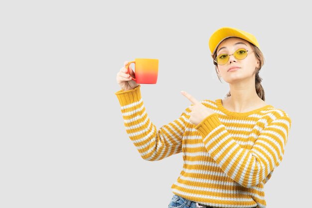 Image of cute young woman in glasses standing and pointing at a mug . High quality photo