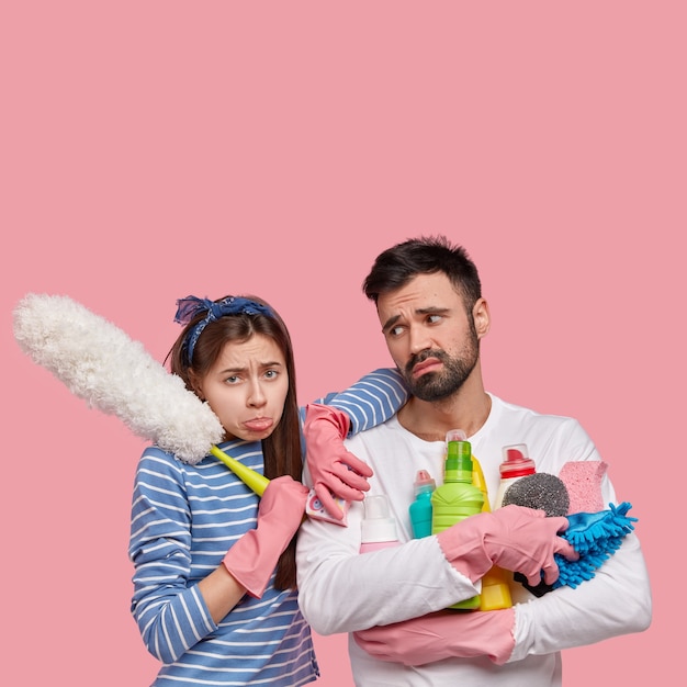 Image of dejected wife and husband clean at home, do spring cleaning, feel fatigue, stand closely to each other, hold detergents