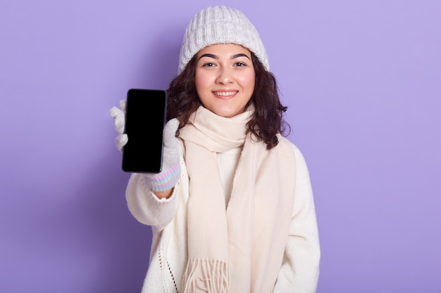 Image of delighted attractive model holding her switched off smartphone in one hand, showing it, blank screen, being in good mood, looking directly at camera, isolated on lilac.