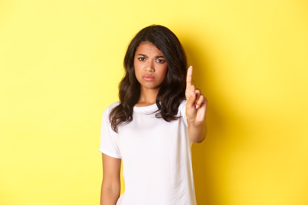 Изображение разочарованной афро-американской девушки, говорящей «нет», встряхивающей пальцем, чтобы запретить или остановить кого-то, не соглашаться с человеком, стоя на желтом фоне.